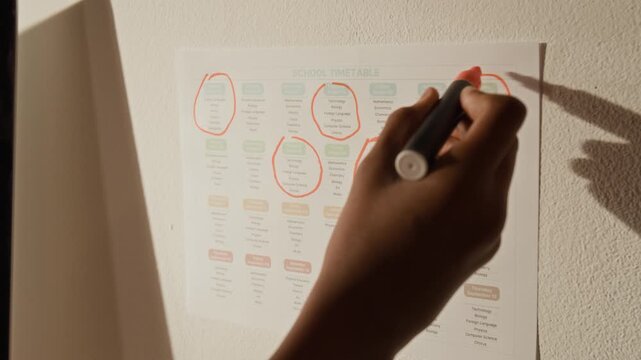 Close-up of hand of disciplined anonymous school student highlighting study days on monthly school timetable, while planning learning activities and setting goals to achieve academic success