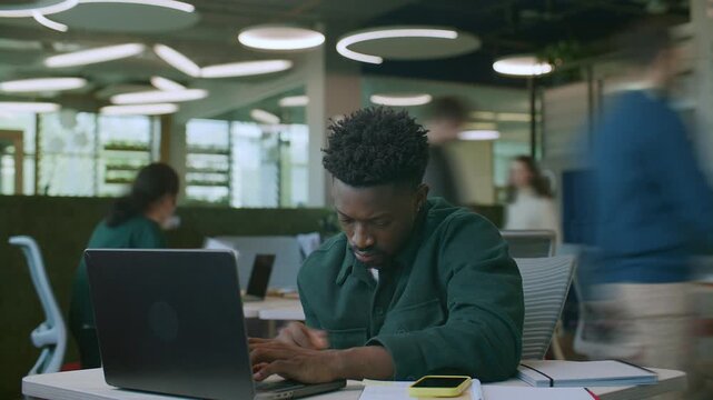 Time lapse of young Black male employee typing on laptop and taking notes at desk during day in corporate office, colleagues working behind him