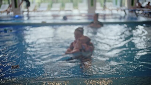 View of an indoor swimming pool where an instructor supports a baby floating on the water during a swimming lesson