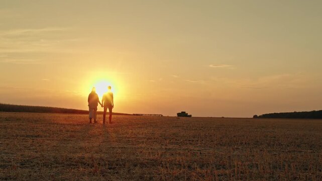 Silhouettes of a couple holding hands walking through a harvested field during sunset.