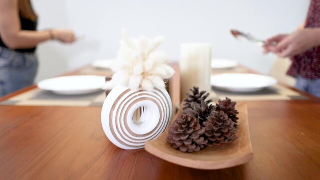 Two Women Setting a Minimalist Dining Table with Pine Cone Centerpiece