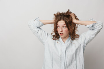 Naklejka premium Surprised young woman with curly hair in white shirt holds head with hands, looking to side. Concept of shock, confusion, and unexpected news in casual studio setting.