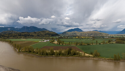 Obraz premium Aerial View Of Fraser Valley River, Farmland And Mountains Under Dramatic Cloudy Sky In British Columbia