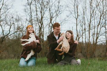 Fototapeta premium Three young adults enjoy a casual outdoor moment holding baby goats in a grassy field with bare trees in the background during a cool season.