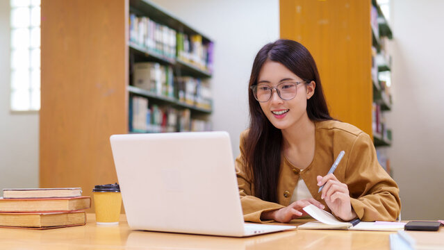 young Asian businesswoman entrepreneur student working on laptop computer taking notes writing report in modern library office space with books background
