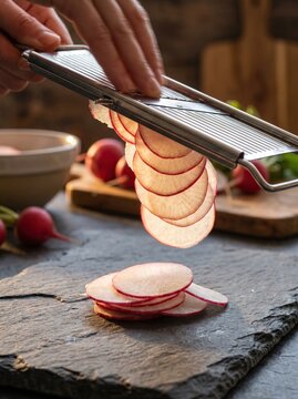 Close up of hand slicing fresh radishes with a mandoline on rustic stone surface in a kitchen