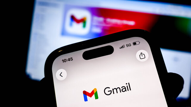 Closeup of a smartphone screen displaying the gmail app logo, with a blurred background showing a computer monitor