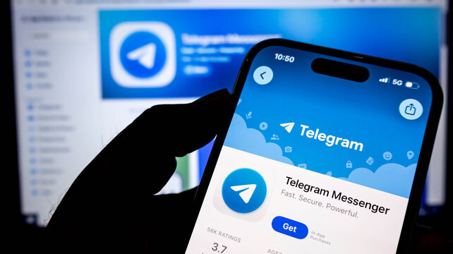 Closeup of a hand holding a smartphone displaying the telegram messenger app page on the apple app store, with a blurred computer screen in the background
