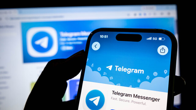 Closeup of a hand holding a smartphone displaying the telegram messenger app store page with a blurred computer screen in the background