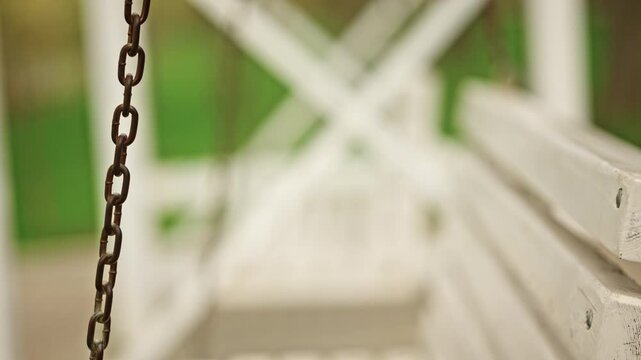 Extreme close up of a metal chain attached to a white wooden swing bench, showing rust details and texture on the metal ring and links