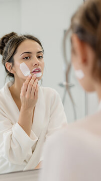 Woman removing mouth tape with cotton pad, bathroom mirror reflection. A woman in a white robe uses a cotton pad to gently remove mouth tape while looking at her reflection in a bathroom mirror.