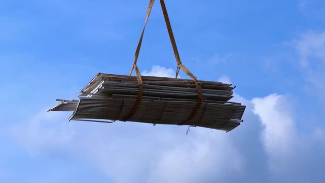 Bundle of long wooden plank wrapped with palm fiber strap hanging suspended midair against blue sky with white cloud, construction lift scene