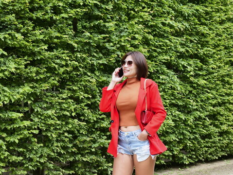 Attractive Asian woman in a red jacket and denim cutoffs smiles while making a phone call by a green hedge