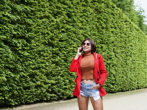 Attractive Asian woman in a red jacket and denim cutoffs smiles while making a phone call by a green hedge