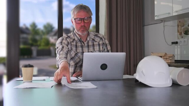Engineer reviewing blueprints and making corrections at home office desk with laptop, construction helmet and technical documents during remote architectural planning and project management work.