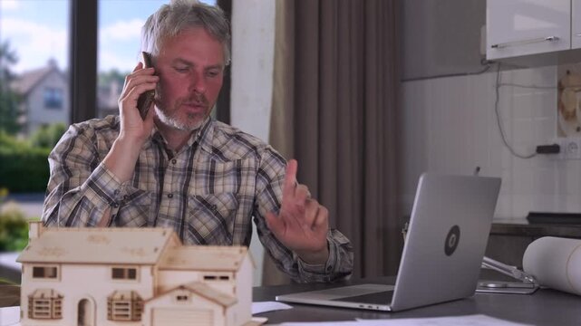 Engineer talking on phone at desk, working remotely from home office with architectural house models and laptop during planning and construction management tasks.