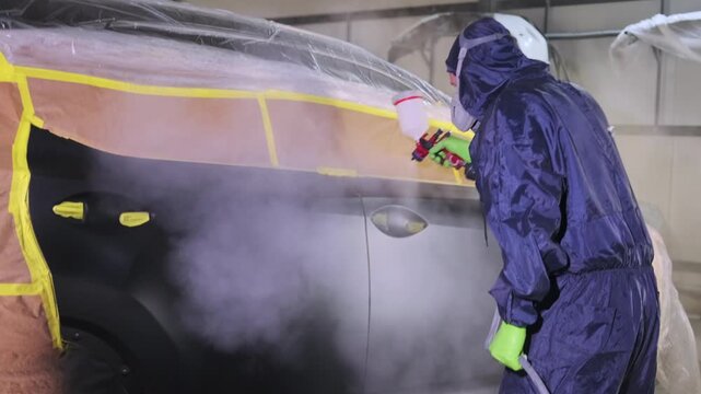 Auto body painter applying glossy clear coat on masked vehicle panel with spray gun, performing final lacquer coverage after collision repair and insurance refinishing service.