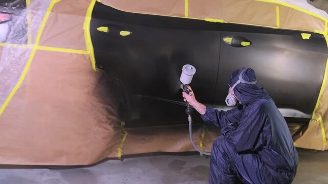 Auto body painter spraying black paint on insured vehicle panel in repair workshop, restoring collision damage during insurance claim refinishing and compensation service