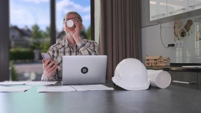 Engineer talking on phone while working remotely at home office desk with laptop, blueprints and construction helmet during online project coordination, planning review and consulting communication.