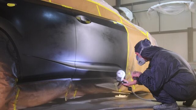 Painter applying final black coating to lower car door with spray gun in bodyshop, completing insurance refinishing and repair compensation workflow after accident damage before quality approval.