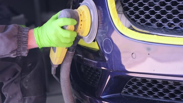 Worker polishing car bumper with orbital machine in auto body shop close-up, detailing damaged surface after insurance claim repair and express maintenance service