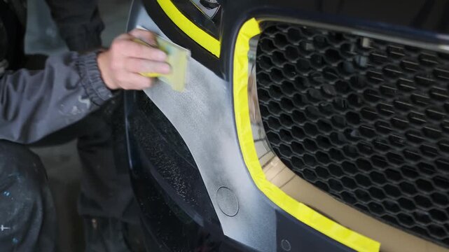 Auto body worker hand sanding front bumper edge beside grille in repair workshop close-up, preparing scratched car surface for repainting, insurance repair and collision restoration service.
