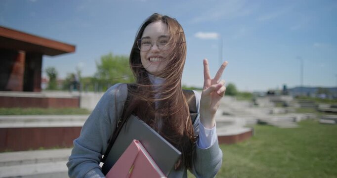 A hand-held portrait of a young female student in casual clothes, wearing glasses and a backpack, holding a notebook and laptop in her hands and making a welcoming gesture with her hands - victory
