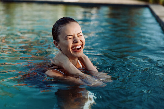 Happy woman swimming in a pool smiling with closed eyes enjoying summer lifestyle and relaxation outdoors on a sunny day with clear blue water surrounding her.