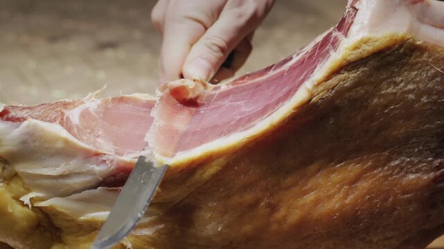 Close up video of a person expertly slicing thin, delicate pieces of prosciutto from a large, cured leg of ham on a rustic wooden surface.