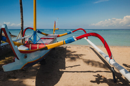 Jukung or kano - traditional fishing boat on Sanur beach in Bali beside calm tropical sea beneath bright blue sky. Scenic island coastline, seaside travel and Indonesian coastal lifestyle