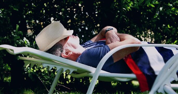 Senior man napping on garden lounge chair with arms crossed and hat covering face, relaxing in shade outdoors during sunny day in peaceful backyard environment