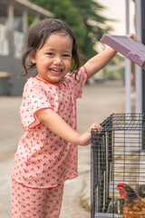 Joyful Child Opening a Chicken Coop A smiling young Southeast Asian girl in cherry-print pajamas happily lifts the lid of a wire cage to interact with a brown hen. © Andi