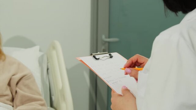 Doctor writes medical notes on clipboard during bedside consultation. Medical specialist records patient complaints updating treatment chart