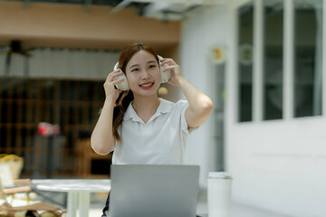 Fototapeta premium Young Asian woman wearing white polo shirt, adjusting wireless headphones, listening to music or podcast while working on laptop outdoors