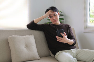 Obraz premium Young man sitting on sofa, holding smartphone, looking upset and having a headache. Experiencing digital stress and frustration