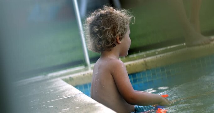 Toddler sitting in shallow pool water by edge with orange toy sticks, smiling and playing joyfully behind visible safety fence during sunny backyard moment