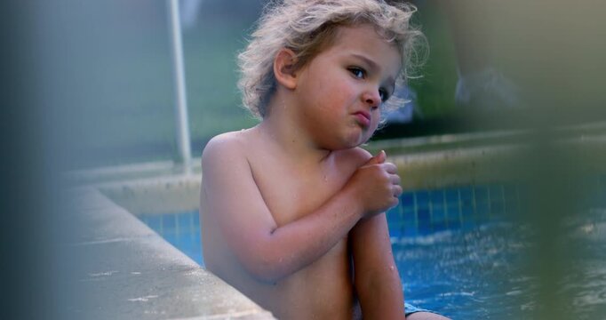 Toddler standing by edge of swimming pool with arms crossed over chest, looking away in quiet thoughtful moment behind safety fence during summer day