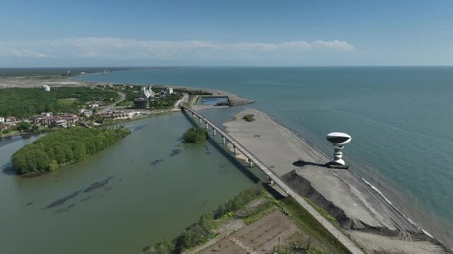 Aerial view of Anaklia resort in Georgia featuring the futuristic observation tower, pedestrian bridge over Enguri river, sandy beach, and Black Sea coastline on a sunny day.