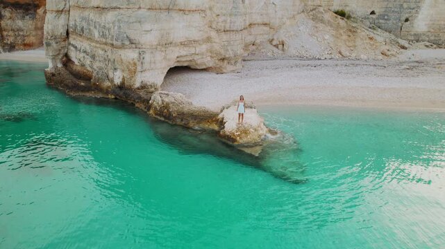 Traveler woman walking on offshore rock facing secluded turquoise cove and white cliffs on Fteri beach in Kefalonia Greece