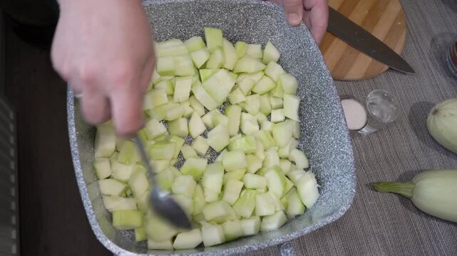A woman stirs sliced ​​zucchini in a deep baking dish. A woman's hand uses a spoon to stir pieces of fresh peeled zucchini in a gray granite mold or roasting pan