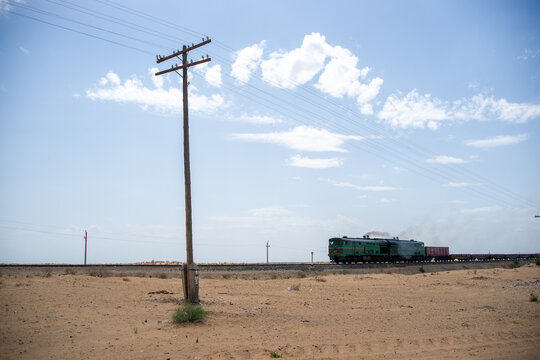 A green freight locomotive moves along distant tracks across a barren desert plain under a vast blue sky with scattered clouds, emphasising isolation, industry and wide open landscapes. Uzbekistan.