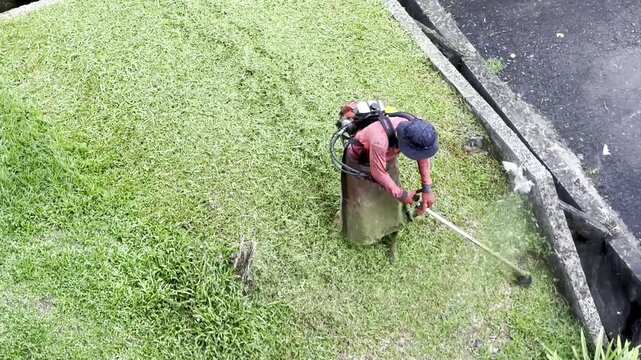 Aerial angle shot of a municipal landscaper using a petrol brush cutter to clear a grassy slope next to a drainage ditch. The footage showcases professional lawn care, heavy-duty trimming.