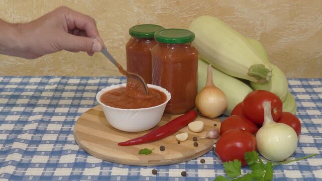 Homemade zucchini caviar in jars and deep bowl in kitchen. Hand scooping spoon with homemade zucchini caviar over deep white bowl filled with sauce