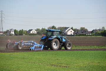 Obraz premium Blue Tractor with Harrow Working on Field Under Sunny Sky with Contrast of Green and Plowed Soil