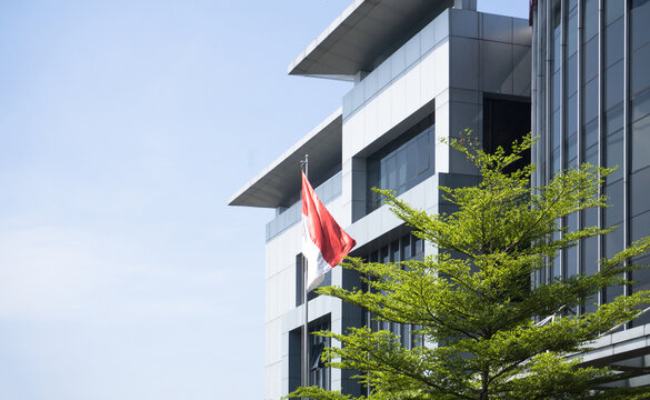 low angle view of the indonesian national flag waving in front of a modern office building with silver metallic cladding and green trees against a clear blue sky