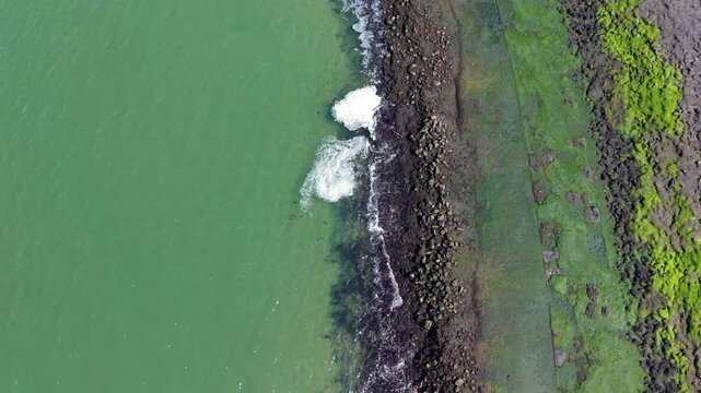 Top down aerial view of a rocky shoreline with green moss and seaweed. Turquoise sea water with white foam crashing against dark stones on the Dutch coast. Natural abstract texture and patterns.