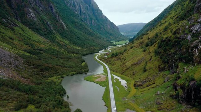 Aerial drone zoom in footage shows vehicles driving on scenic valley road alongside flowing river in Klakegg, Norway. Majestic green mountains landscape captured during sunny July summer day
