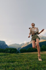 Fototapeta premium Young woman strolls across a sunlit alpine meadow with distant mountains, feeling freedom and joy as she moves in hiking boots under a clear sky.