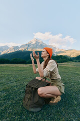 Fototapeta premium Woman kneeling in a grassy field with distant mountains, wearing an orange beanie and backpack. A candid outdoor lifestyle shot celebrating nature, exploration, and adventure.