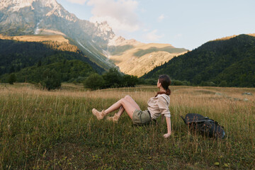 Fototapeta premium A woman rests in a sunlit meadow with distant mountains, backpack beside her. A calm moment of outdoor life, hiking, and nature, inviting exploration and solitude.